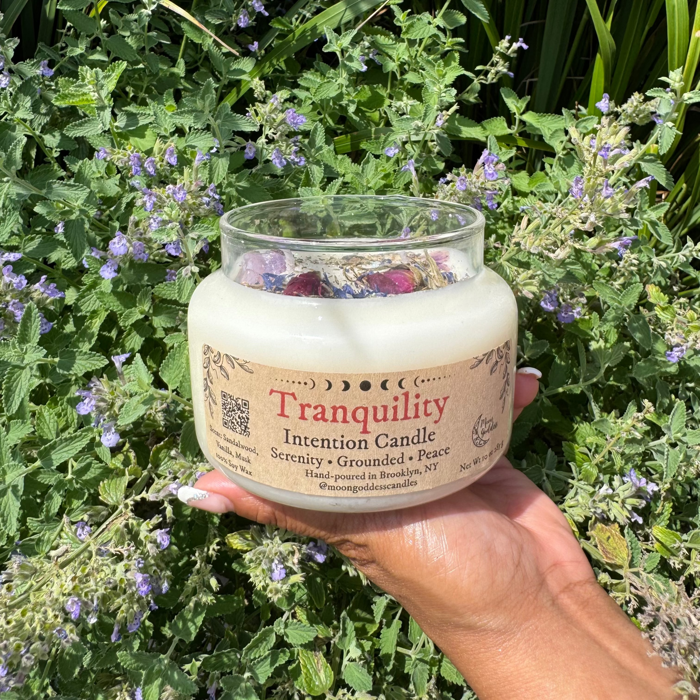 Hand holding a 'Tranquility' intention candle in front of green foliage