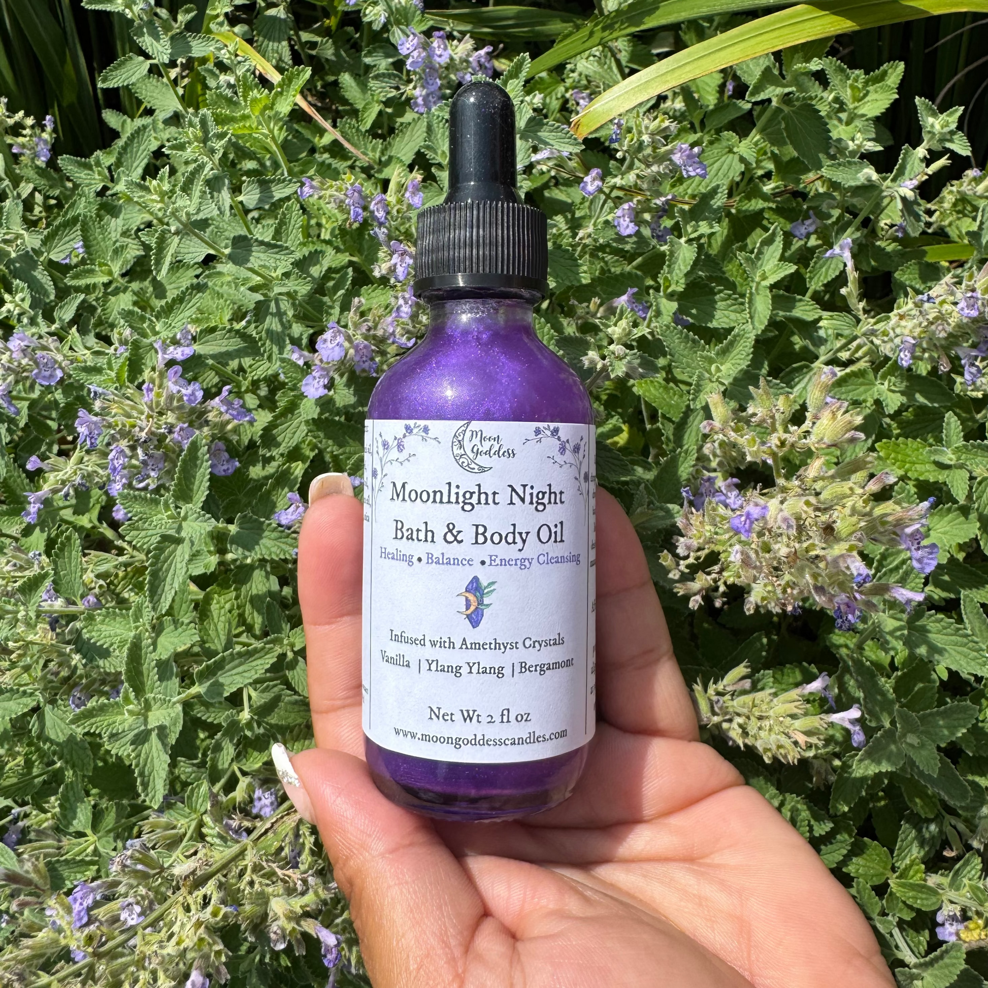 Moonlight Night Bath & Body Oil by Moon Goddess Candles, a shimmering purple oil infused with amethyst crystals, vanilla, ylang ylang, and bergamot, held in hand against a backdrop of blooming purple flowers for healing, balance, and energy cleansing