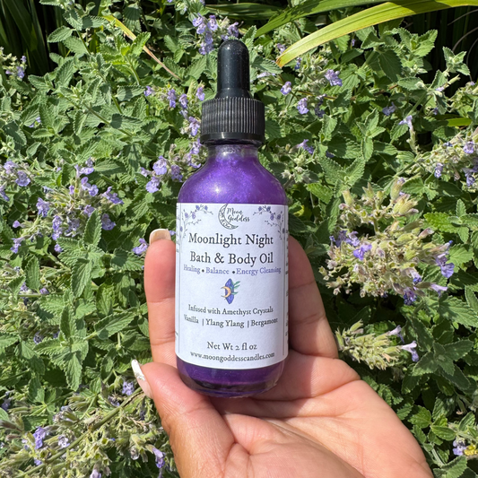 Moonlight Night Bath & Body Oil by Moon Goddess Candles, a shimmering purple oil infused with amethyst crystals, vanilla, ylang ylang, and bergamot, held in hand against a backdrop of blooming purple flowers for healing, balance, and energy cleansing