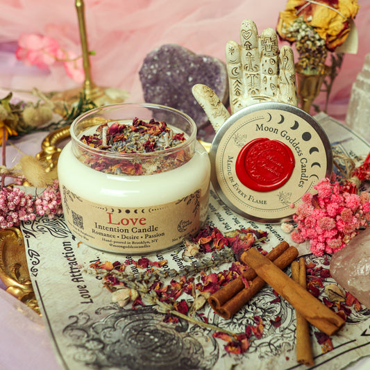 Love Intention Candle by Moon Goddess Candles, hand-poured in Brooklyn and topped with rose petals and herbs, surrounded by cinnamon sticks, crystals, and pink floral decor to inspire romance, passion, and desire
