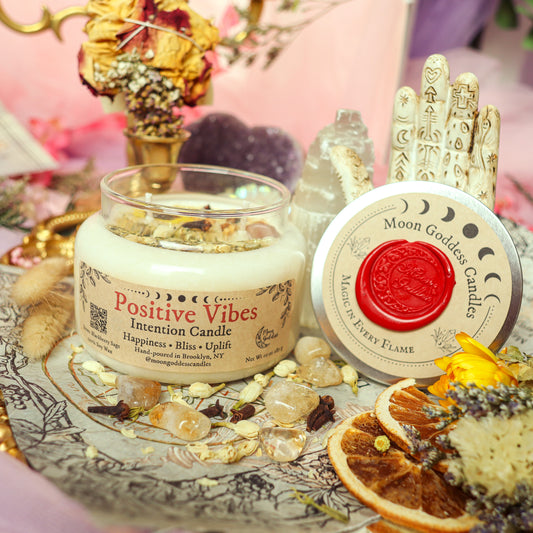Positive Vibes Intention Candle by Moon Goddess Candles, hand-poured in Brooklyn and infused with herbs and crystals, surrounded by dried florals, orange slices, and spiritual decor on a mystical altar setup